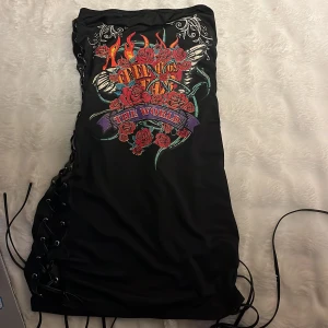 Svart klänning med tryck och snörning - Cool svart klänning med  tryck av rosor och eldflammor samt texten 'The World'. Klänningen har en unik snörningsdetalj på båda sidorna som ger en edgy look.