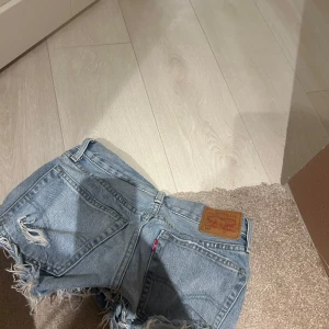 Blå jeansshorts med slitningar - Säljer ett par coola blå jeansshorts från Levi's med slitna detaljer och fransar. Perfekta för en avslappnad stil. De har en klassisk femficksdesign och knappgylf.