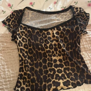 Leopardmönstrad topp med spetsdetaljer - Snygg leopardmönstrad topp med spetsdetaljer vid halsringningen. Toppen har korta ärmar och en elegant design som passar perfekt för en trendig look.