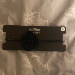 Svart halsband med ros från Ur&Penn - Elegant svart halsband från Ur&Penn med en vacker rosdetalj i mitten. Halsbandet har svarta pärlor och en justerbar kedja i guld för perfekt passform. Perfekt för att ge en stilfull touch till din outfit. Aldrig använd och helt ny köpt. 