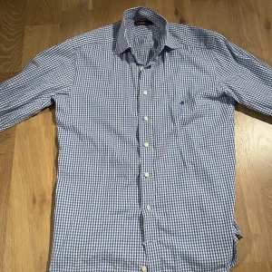 Blå och vit rutig skjorta från Morris - Snygg blå och vit rutig skjorta från Morris . Skjortan har ett diskret broderi på bröstet och är tillverkad i bomull. Perfekt för en stilren look! Skjortan är storlek S och passar ca 160-175. Modellen är 170cm 65kg