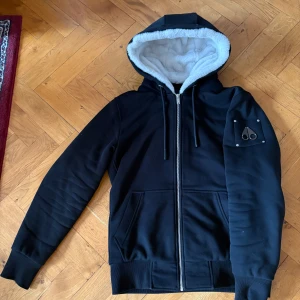 Svart hoodie från Moose Knuckles - Säljer en svart hoodie från Moose Knuckles med dragkedja och vit fleecefoder i luvan. Jackan har ett unikt märke på ärmen och ribbade muddar. Perfekt för kyliga dagar.