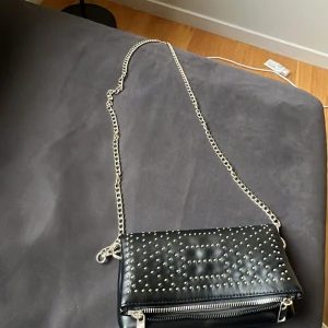 Svart axelväska från Even&Odd - Snygg svart axelväska från Even&Odd med nitar och kedja. Väskan har en dragkedja och magnetknappar för säker förslutning. Perfekt för att ge en edgy touch till din outfit.