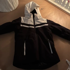 Svart och vit jacka med huva - Snygg svart och vit jacka med huva och dragkedja framtill. Jackan har långa ärmar och två fickor med dragkedjor. Perfekt för kyligare dagar passar super bra till skidåkning då man inte behöver ha så mycket kläder under + regniga och blåsiga dagar.