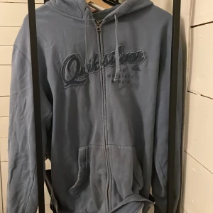 Blå hoodie från Quiksilver - Säljer en blå hoodie från Quiksilver med dragkedja och broderad logga på bröstet. Perfekt för en avslappnad stil med sina långa ärmar och bekväma passform. Huvan har justerbara snören för extra komfort.