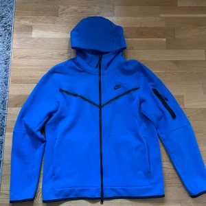 Blå hoodie från Nike - Säljer en blå hoodie från Nike med dragkedja och huva. Jackan har svarta detaljer och en ficka på ärmen. Perfekt för en sportig look.