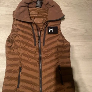 Brun dunväst från lars Mønsen - Snygg brun dunväst från Lars Mønsen med dragkedja och hög krage. Perfekt för kyliga dagar och har en stilren design med ett litet märke på bröstet. Västen är lätt och bekväm att bära. Helt ny, köpte den för 1250 kronor