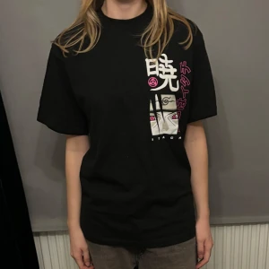 Svart t-shirt med anime-tryck - Cool svart t-shirt med stort anime-tryck fram och bak. Designen har vita och rosa detaljer med japanska tecken och en karaktärsbild. Perfekt för anime-fans som vill ha en unik stil.