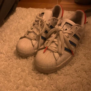 Adidas Superstar sneakers - Klassiska Adidas Superstar sneakers i vitt med blå och röda detaljer. Skorna har snörning och den ikoniska tåhättan. Baktill finns en röd detalj med Adidas-loggan.