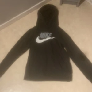 Svart hoodie från Nike - Säljer en svart hoodie från Nike med det klassiska logotyptrycket i vitt på bröstet. Tröjan har en bekväm huva och långa ärmar, perfekt för en avslappnad stil.