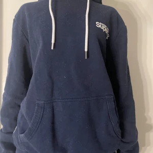 Mörkblå hoodie från Superdry - Säljer en mörkblå hoodie från Superdry med vit logga på bröstet och justerbara snören i huvan. Tröjan har en stor ficka framtill och en blommig insida i huvan. Perfekt för en avslappnad stil. NYPRIS 1500 vårt 250