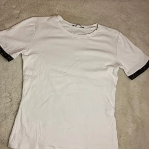 Vit t-shirt från Zara med glittriga ärmslut - Säljer en stilren vit t-shirt från Zara med korta ärmar och glittriga detaljer vid ärmsluten. Perfekt för att ge en touch av glamour till din vardagslook. Tillverkad i mjukt material för en bekväm passform.