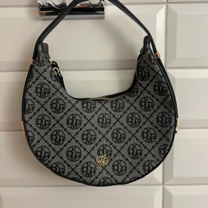 Svart och beige axelväska med SHEIN eller Temu  - Snygg axelväska i svart och beige med monogrammönster från Ralph Lauren. Väskan har en dragkedja upptill och guldfärgade detaljer. Perfekt för att ge din outfit en lyxig touch. Aldrig använd. Säljer för 100 kronor. 