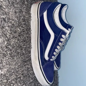 Blå och vita sneakers från Vans - Snygga blå sneakers från Vans med vita detaljer och klassisk snörning. Perfekta för en avslappnad stil. Skorna har en vit sula och den ikoniska Vans-loggan på hälen.