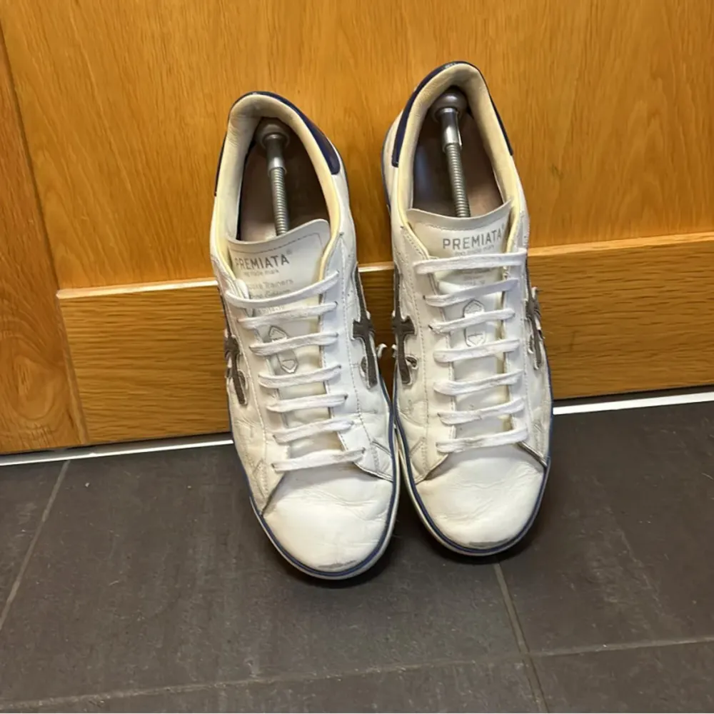 Snygga vita sneakers från Premiata med mörkblå detaljer och en unik svart stjärndesign på sidorna. Skorna har snörning och en klassisk siluett som passar perfekt för en avslappnad stil.. Kengät.