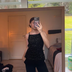 Svart glittrig topp - Säljer en snygg svart glittrig topp med smala axelband och öppen rygg. Perfekt för en kväll ute eller festliga tillfällen. Toppen har en figurnära passform och ger en elegant look. Slutsåld på Gina 