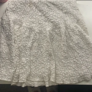 Vit kjol med struktur - Säljer en vit kjol med en unik strukturerad yta. Kjolen har en lätt volang och är perfekt för en stilren look. Den är kort och passar bra till olika tillfällen. Perfekt för dig som vill ha något annorlunda i garderoben!