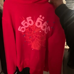 Röd Sp5der hoodie - Säljer en cool röd hoodie med unikt tryck i vitt och blått. Hoodien har långa ärmar och en klassisk passform. Perfekt för en avslappnad stil.