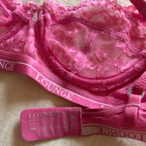 Rosa spets-BH från Lounge - Säljer en vacker rosa spets-BH från Lounge med blommigt mönster. Den har justerbara axelband och en elastisk kant med Lounge-loggan. Perfekt för en bekväm och stilren look.