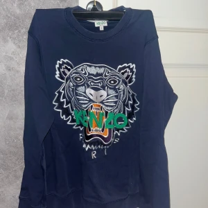 Mörkblå sweatshirt från Kenzo - Säljer en mörkblå sweatshirt från Kenzo med ett stort tigerhuvud och texten 'Kenzo Paris' broderat på framsidan. Tröjan har långa ärmar och en rund halsringning. Perfekt för en cool och avslappnad stil. Tröjan är som ny och skulle sätta skicket som 9/10 med tanke på vardaglig användning😊