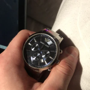 Emporio Armani klocka - Snygg Emporio Armani klocka med en stilren svart urtavla och tre subdialer. Boetten och armbandet är i silverfärgat rostfritt stål. Perfekt för den som vill ha en elegant och tidlös accessoar.      Herrur Emporio Armani, stål, quartz, ca 43mm, ref nr: AR-2448, snr: 111204, stållänk, innermått ca 18cm, väldigt fint skick, finns ej låda eller kvitto. Pris kan diskuteras vid snabb affär