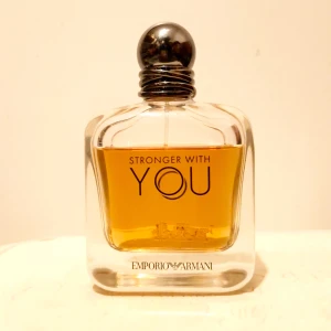 Stronger With You parfym från Emporio Armani - Säljer en elegant parfymflaska av Emporio Armani, 'Stronger With You'. Flaskan har en stilren design med en rundad svart kork och en klar glasflaska som visar den gyllene doften inuti. Perfekt för den som vill ha en sofistikerad och modern doft. Garanterat att ge en komplimanger. Runt 90ml 