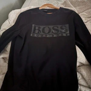 Svart tröja från Hugo Boss - Säljer en stilren svart tröja från Hugo Boss med glittrig logga på bröstet. Tröjan har långa ärmar och en klassisk rund hals. Lite slitage vid armbågarna. 