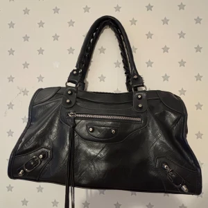 Svart skinnväska 2000s bag y2k - Snygg svart skinnväska med flätade handtag och coola metalldetaljer. Väskan har en dragkedja framtill och flera nitar som ger en edgy look. Perfekt för att bära med sig allt man behöver med stil.