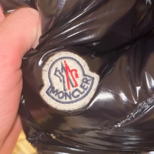 Svart dunjacka från Moncler - Säljer en svart glansig dunjacka från Moncler med klassisk logga på bröstet. Jackan har en quiltad design och är perfekt för kyligare väder. Inuti finns en unik serietidningsdetalj som ger en cool touch. Perfekt för dig som vill ha stil och funktion i ett!