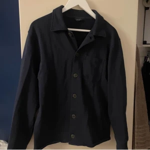 Mörkblå skjorta från Riley - Snygg mörkblå skjorta från Riley med klassisk krage och knappar framtill. Skjortan har en bröstficka och långa ärmar, perfekt för en stilren look. Passar bra till både jeans och chinos. Storlek M och är använd 1 gång, den är även jätte true to size 