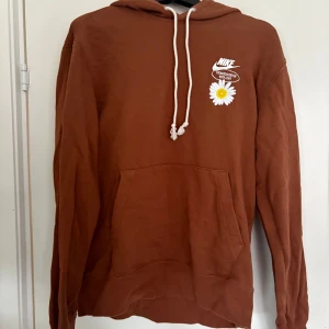Brun hoodie från Nike med blommotiv - Säljer en brun hoodie från Nike med en broderad prästkrage och texten 'From Beaverton With Love' på framsidan. Baksidan har ett stort tryck med solrosor och texten 'Have a Nike Day'. Perfekt för en avslappnad stil! Köpt i Paris för ca 1000kr, tror inte det gick art köpa den online, så ganska orginell.