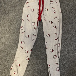Grå mjukisbyxor med Hello Kitty-mönster - Säljer ett par grå mjukisbyxor med ett gulligt Hello Kitty-mönster. Byxorna har en röd snörning i midjan för justerbar passform. Perfekta för en avslappnad dag hemma.