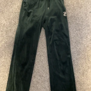 Gröna mjukisbyxor från Juicy Couture - Snygga gröna mjukisbyxor från Juicy Couture med glittrig text på baksidan. Byxorna har en bekväm passform med dragsko i midjan och är perfekta för en avslappnad stil. Märkesdetaljer på snöret ger en extra touch.