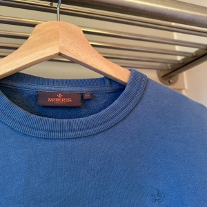 Blå fräsch tröja från Morris - Säljer en stilren blå tröja från Morris. Tröjan har en diskret logga broderad på bröstet och en etikett nedtill. Perfekt för en avslappnad look. Ny pris: 1399 kr