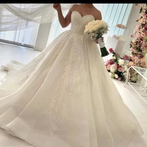 Vit brudklänning med spetsdetaljer - Elegant vit brudklänning med vackra spetsdetaljer och en voluminös kjol. Klänningen är axelbandslös och har en klassisk, romantisk stil som passar perfekt för ett bröllop.