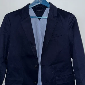 Svart kavaj från Tommy Hilfiger - Snygg svart kavaj från Tommy Hilfiger med klassisk design. Kavajen har två knappar framtill och en bröstficka. Perfekt för både formella och semi-formella tillfällen. Storlek 176, Färg Mörk Blå
