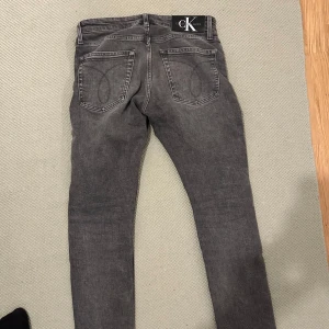 Grå slim jeans från Calvin Klein - Snygga grå slim jeans från Calvin Klein med klassisk femficksdesign och knappgylf. Perfekta för en stilren look.