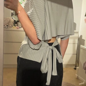 Plisserad topp med öppen rygg och knytband - Säljer en super snygg stilren grågrön plisserad topp med korta ärmar och knytband i ryggen. Köpte för ett år sen på Gina Tricot o använd några gånger men inga direkta tecken på användning!