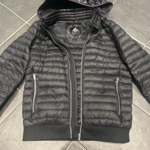 Svart quiltad jacka från Moose Knuckles - Säljer en svart quiltad jacka från Moose Knuckles med dragkedja och huva. Jackan har en avtagbar luva och två fickor med dragkedja framtill. Perfekt för kyligare dagar med sin stilrena design och funktionella detaljer.