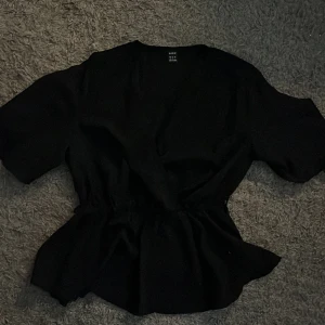Svart blus från Shein - Säljer en stilren svart blus från Shein i storlek XS. Blusen har korta ärmar och ett medföljande bälte för att framhäva midjan. Perfekt för en elegant look.