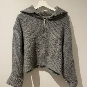 Grå stickad tröja med dragkedja - Mysig grå stickad tröja med dragkedja framtill och en bred krage. Tröjan har långa ärmar och en avslappnad passform, perfekt för kyligare dagar.