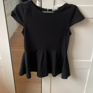 Svart topp med volang  - Snygg svart peplumtopp med korta ärmar och dragkedja i ryggen. Perfekt för en stilren look med en feminin touch. Toppen har en figursydd passform och är tillverkad i ett mjukt material.