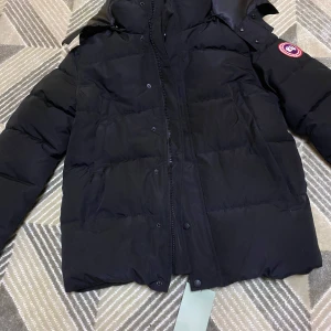 Svart dunjacka från Canada Goose - Säljer en svart dunjacka från Canada Goose med en klassisk design och varumärkets logga på ärmen. Jackan har en dragkedja framtill och är perfekt för kalla vinterdagar.