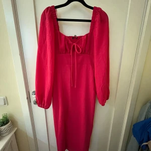Rosa klänning från Gina  - Snygg rosa klänning från gina med långa ärmar och smockdetaljer på ryggen. Klänningen har en söt knytning framtill och en bekväm passform. Perfekt för att sticka ut med sin klara färg och stilrena design. Den har en dragkedja på sidan. Klänningen ser röd ut på bilderna men den är väldigt stark rosa