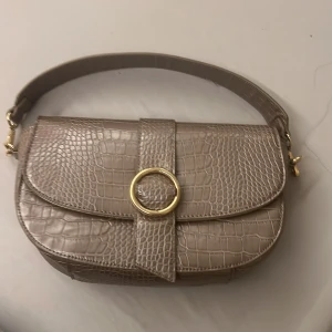 Beige axelväska - Snygg beige axelväska med krokodilmönster och guldfärgad ringdetalj på framsidan. Väskan har en bred axelrem och är perfekt för att ge din outfit en trendig touch. Den är både stilren och praktisk för vardagsbruk.