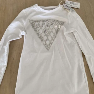 Vit långärmad tröja från Guess Kids - Säljer en stilren vit långärmad tröja från Guess Kids. Tröjan har ett triangulärt mönster med glittriga detaljer på framsidan. Perfekt för en trendig look. storlek 8 år för barn
