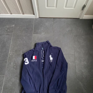 Mörkblå hoodie från Polo Ralph Lauren XL/L - Säljer en snygg mörkblå hoodie från Polo Ralph Lauren med dragkedja och broderade detaljer. Tröjan har nummer 3 och texten 'FRANCE' med en fransk flagga på både fram- och baksidan. Perfekt för en sportig look! Sitter som L