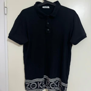 Svart kortärmad pikétröja från Kenzo - Snygg svart pikétröja från Kenzo med knappar och ett unikt mönster längst ner. Perfekt för en stilren look med en touch av Paris. Kortärmad och bekväm passform.