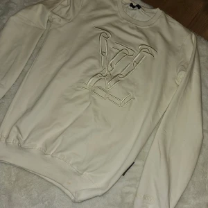 Vit sweatshirt med broderad logotyp - Säljer en stilren vit sweatshirt från Louis Vuitton med en broderad logotyp på framsidan. Tröjan har långa ärmar och en rund halsringning. Perfekt för en avslappnad men ändå lyxig look.