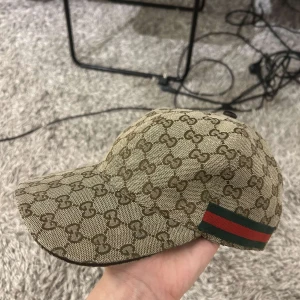 gucci keps - Snygg beige keps från Gucci med klassiskt monogrammönster och röd-gröna detaljer. Perfekt för att ge din outfit en lyxig touch. Tillverkad i Italien.